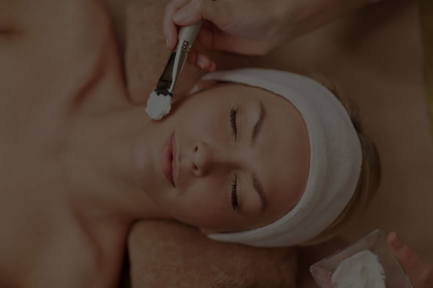 Person lying down with eyes closed, a towel wrapped around their head, receiving a facial treatment.